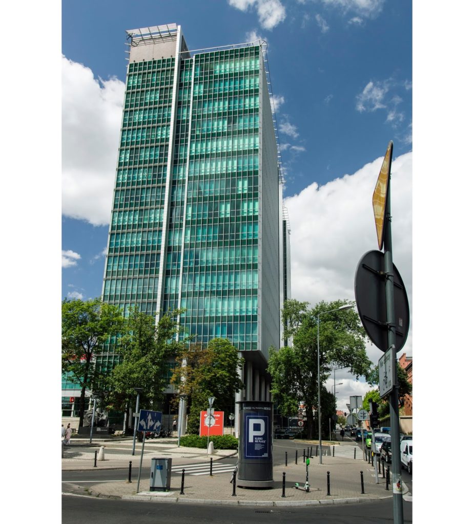 Szklana fasada budynku Financial Centre. Mieniąca się w słońcu na niebiesko tafla szkła z elementami stalowymi. Dominujące pociągłe pionowe linie. Na pierwszym planie słup ogłoszeniowy.