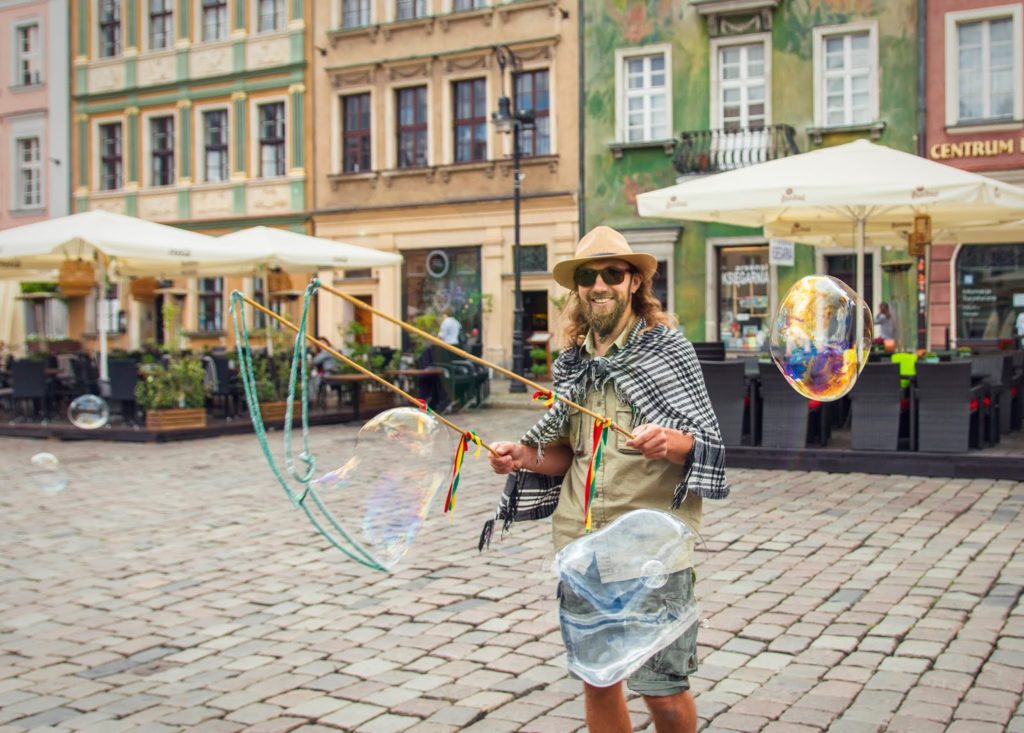 Na zdjęciu widać uśmiechniętego mężczyznę w kapeluszu, który puszcza bańki mydlane. w tle zabytkowe kamieniczki otaczające Stary Rynek w Poznaniu z restauracyjnymi ogródkami