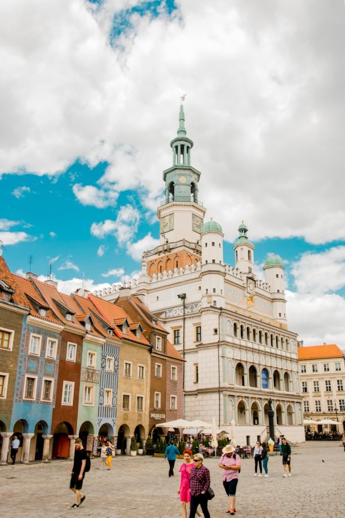 Renesansowy Ratusz stojący na środku poznańskiego Starego Rynku. Po jego lewej stronie kolorowe domki budnicze. Po bruku w cieniu ratusza przechadzają się turyści